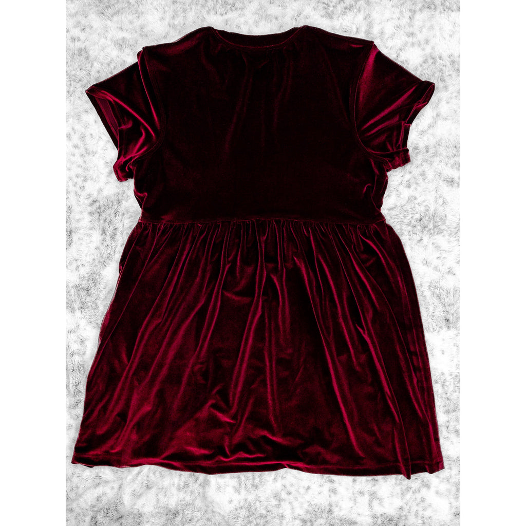 A flat lay image showcasing the back of a wine velvet babydoll dress with short sleeves. The dress features soft, luxurious fabric in rich colors. The background is neutral, highlighting the dress's details and textures.