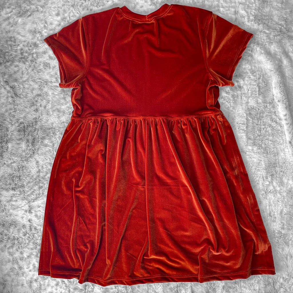 A flat lay image showcasing the back of a rusty orange velvet babydoll dress with short sleeves. The dress features soft, luxurious fabric in rich colors. The background is neutral, highlighting the dress's details and textures.