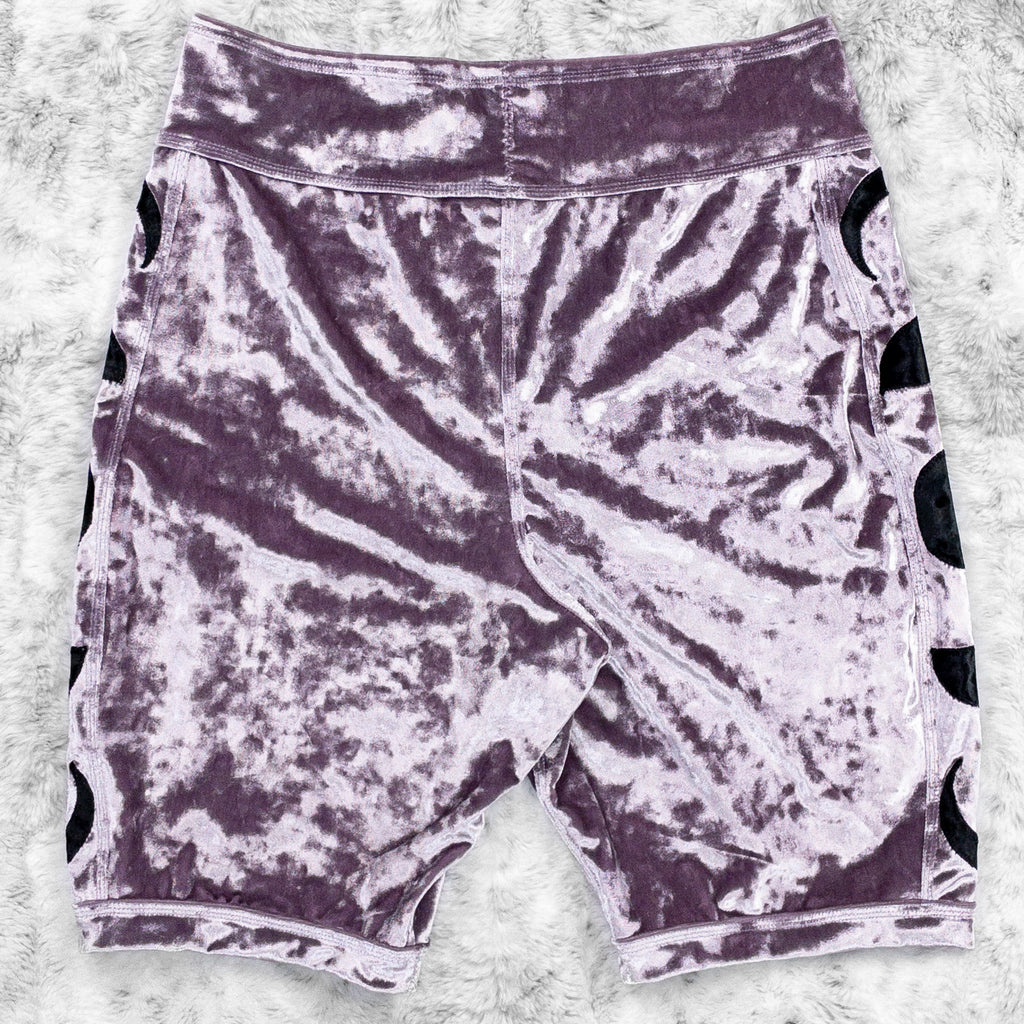 A flat lay image showcasing the back of a purple haze crushed velvet bike shorts with moon phase appliqué along the side seams. The shorts feature soft, luxurious fabric in an icy cool pastel. The background is neutral, highlighting the shorts' details and textures.