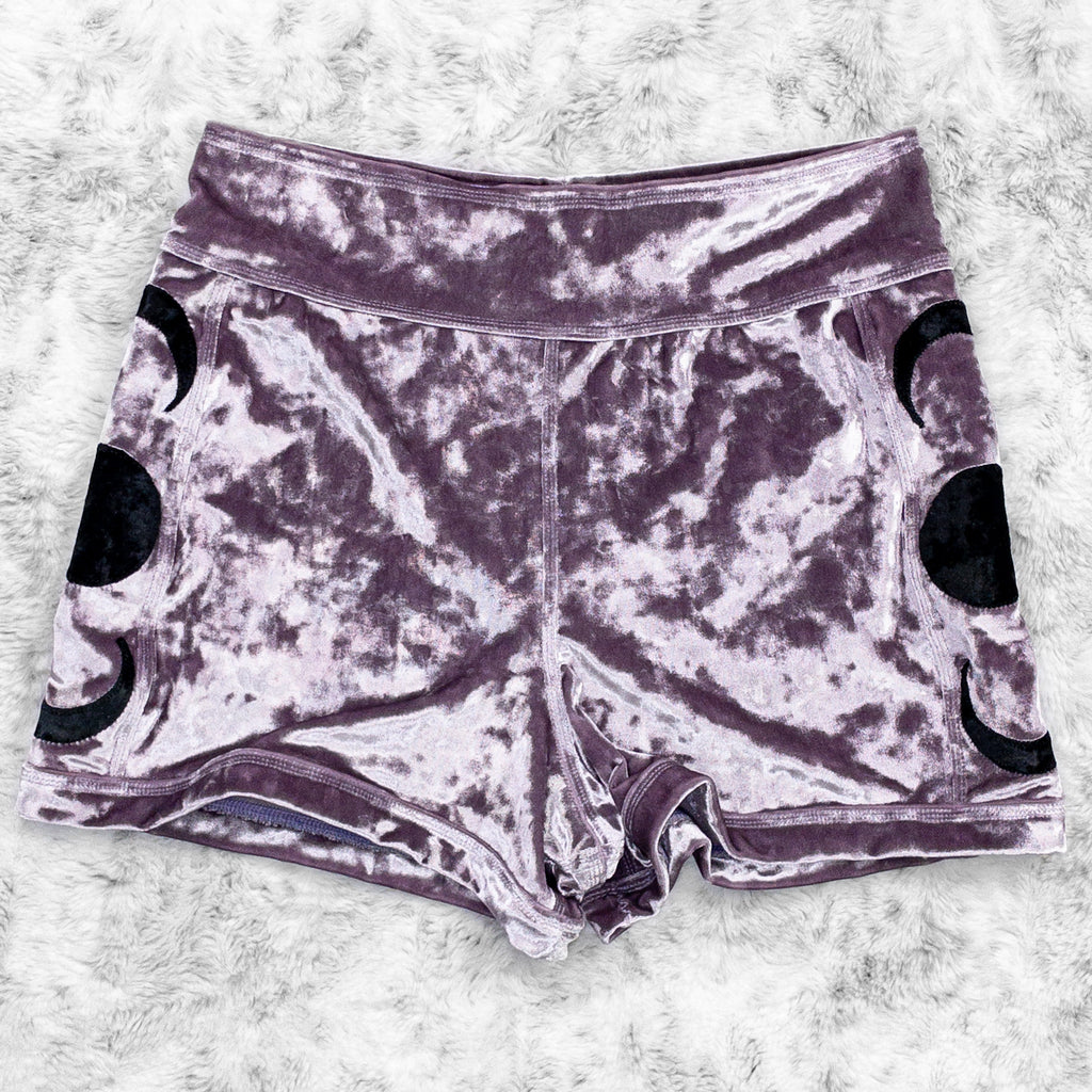 A flat lay image showcasing purple haze crushed velvet booty shorts with moon phase appliqué along the side seams, showcasing a stylish and unique design. The shorts feature soft, luxurious fabric in an icy cool pastel. The background is neutral, highlighting the shorts' details and textures.