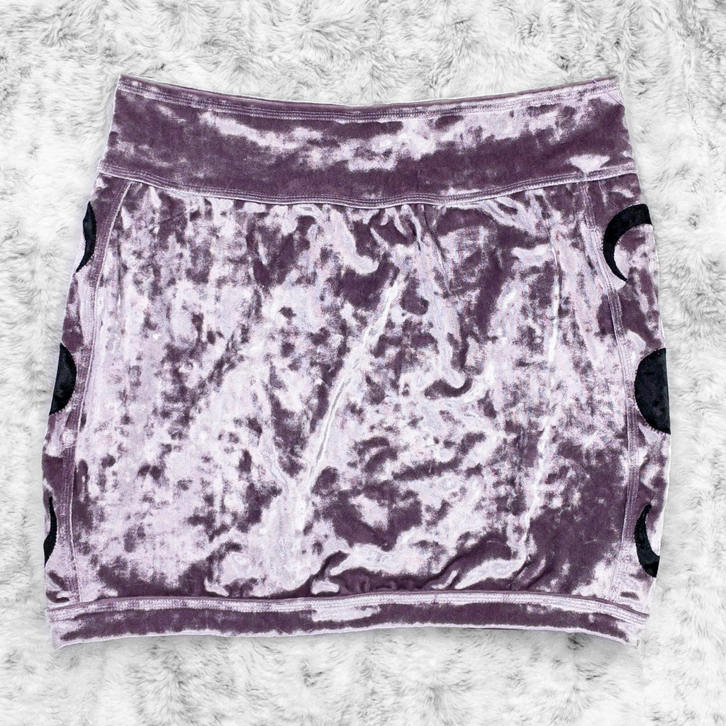 A flat lay image showcasing a purple haze crushed velvet mini skirt with moon phase appliqué along the side seams, showcasing a stylish and unique design. The shorts feature soft, luxurious fabric in an icy cool pastel. The background is neutral, highlighting the shorts' details and textures.