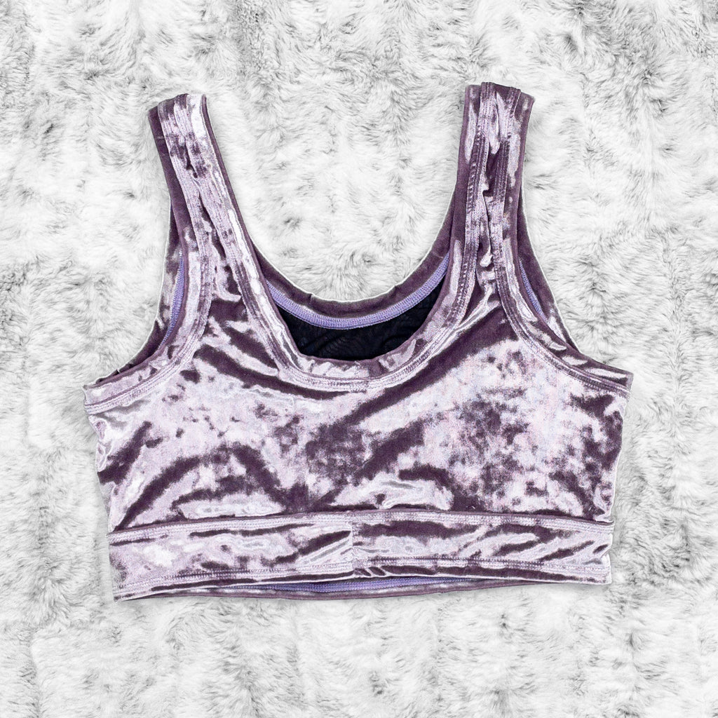 A flat lay image showcasing the back of a purple haze crushed velvet scoopneck close cut crop with princess seams and a crescent moon appliqué on the centerfront. The top features soft, luxurious fabric in an icy cool pastel purple. The background is neutral, highlighting the shorts' details and textures.