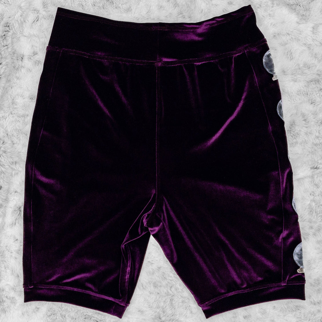 A flat lay image showcasing the back of a plum stretch velvet bike shorts with crystal ball appliqué along the side seams. The shorts feature soft, luxurious fabric in rich colors. The background is neutral, highlighting the shorts' details and textures.