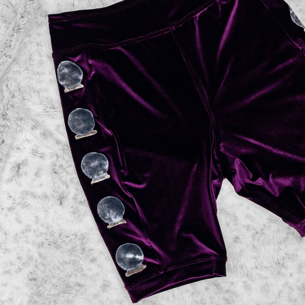 A close up flat lay image showcasing the five hand-cut crystal balls appliqués along the side seam of the plum stretch velvet bike shorts. The shorts feature soft, luxurious fabric in rich colors. The background is neutral, highlighting the shorts' details and textures.