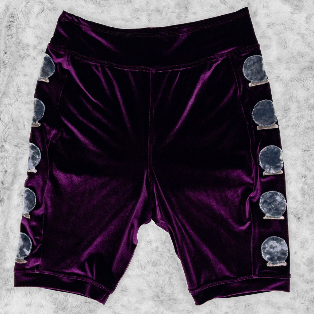 A flat lay image showcasing plum stretch velvet bike shorts with crystal ball appliqué along the side seams, showcasing a stylish and unique design. The shorts feature soft, luxurious fabric in rich colors. The background is neutral, highlighting the shorts' details and textures.