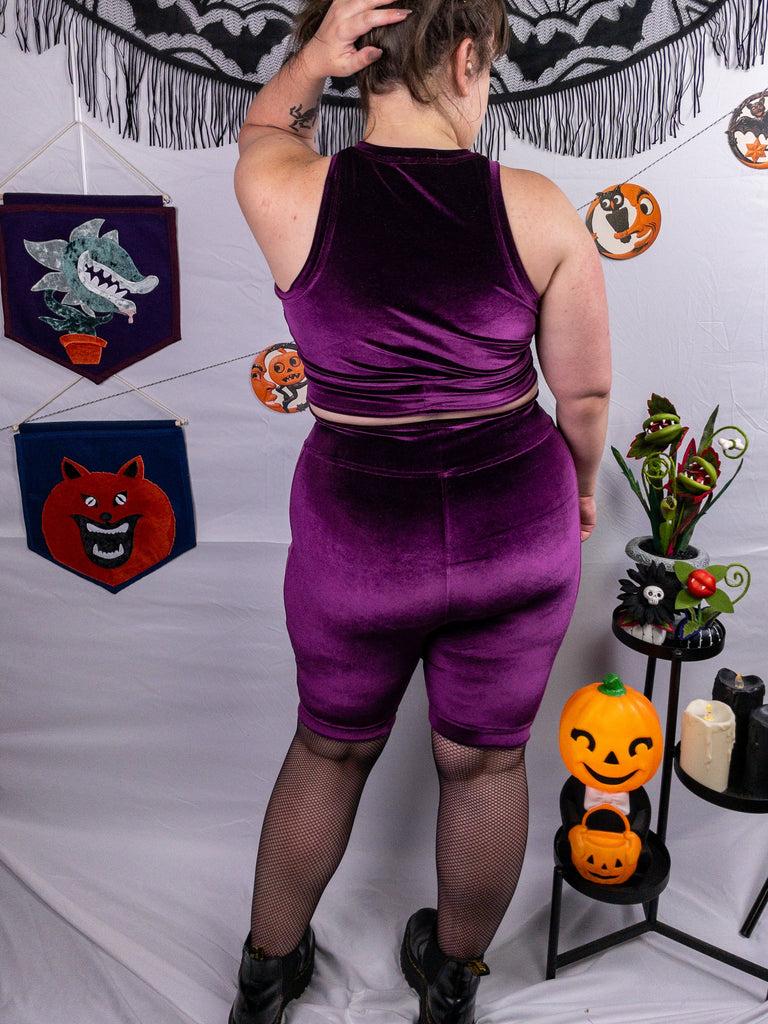 Back view of a mid-size model wearing a plum stretch velvet bike shorts with crystal ball appliqué along the side seams. The photo showcases the rich, deep hue of the fabric. The model stands in front of a pain white background accented with Halloween decorations.
