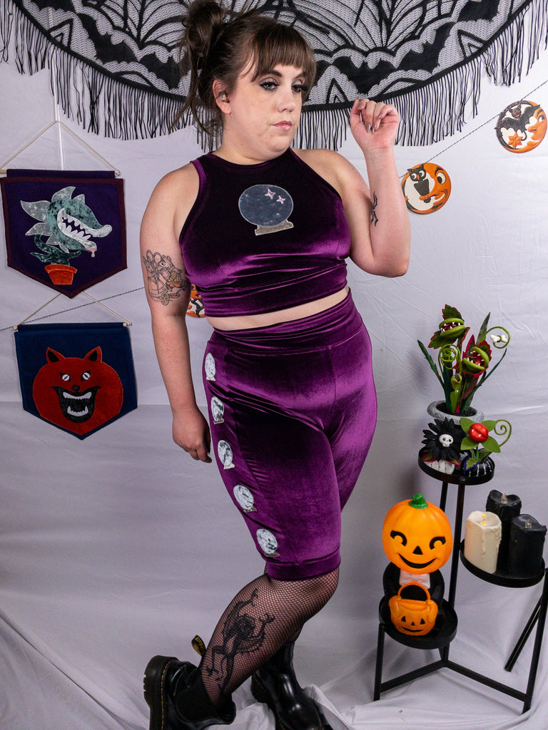 Front side view of a mid-size model wearing a plum stretch velvet bike shorts with five crystal ball appliqués along the side seams and a crewneck close cut crop top out of the same plum stretch velvet as the shorts and a larger crystal ball appliqué. Model is accessorized with black fishnets, and black platform Chelsea boots. The photo showcases the rich, deep hue of the fabric. The model stands in front of a pain white background accented with Halloween decorations.