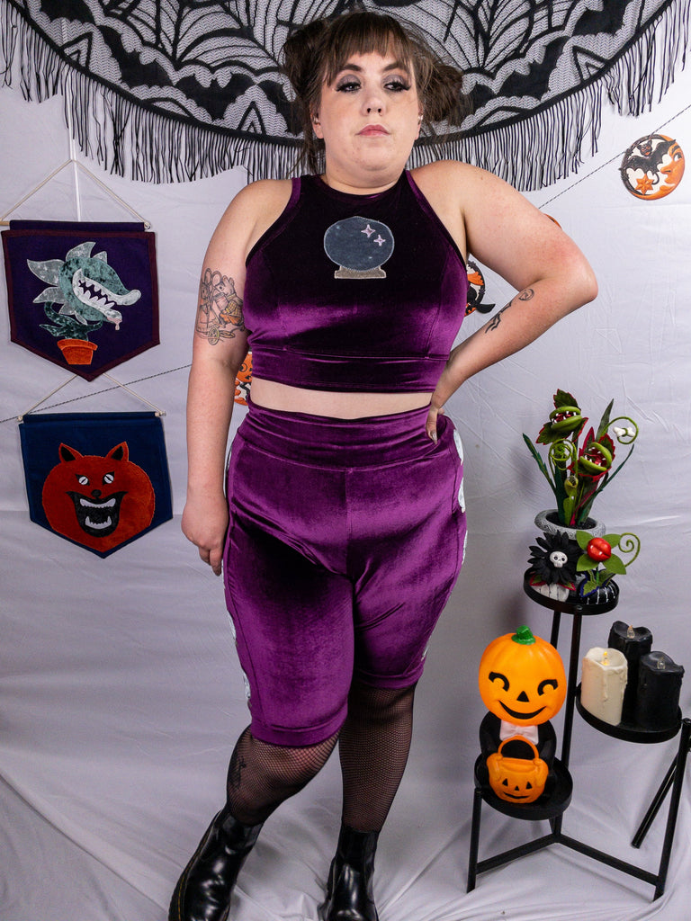 Front view of a mid-size model wearing a plum stretch velvet bike shorts with crystal ball appliqué along the side seams and crewneck close cut crop top out of the same plum stretch velvet as the shorts and a larger crystal ball appliqué. Model is accessorized with black fishnets, and black platform Chelsea boots. The photo showcases the rich, deep hue of the fabric. The model stands in front of a pain white background accented with Halloween decorations.