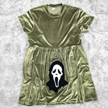 A flat lay image showcasing a olive green velvet babydoll dress with short sleeves adorned with an evocative Ghostface appliqué from 90s meta-horror classic Scream (1996). The dress features soft, luxurious fabric in rich colors. The background is neutral, highlighting the dress's details and textures.