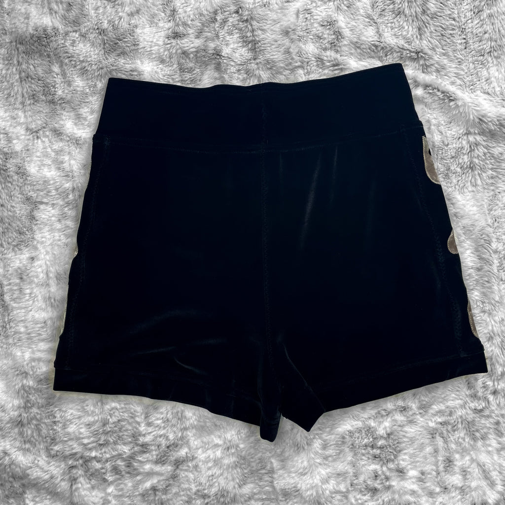 A flat lay image showcasing the back of premium smooth black velvet booty shorts with witchy planchette appliqué along the side seams. The shorts feature soft, luxurious fabric in an inky black. The background is neutral, highlighting the shorts' details and textures.